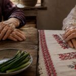 Two pairs of elderly women's hands, one on a wooden Chinese kitchen table, the other on an Albanian embroidered cloth, in warm light