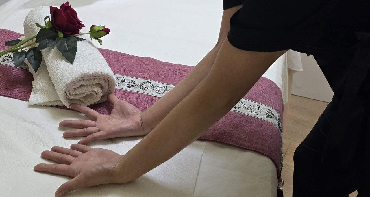 Rolled white towels, smooth river pebbles in a dark ceramic bowl, and a dried chrysanthemum sprig on the massage bed in the warm khaki-olive room at Chinese Massage - Tai Chi Tirana.